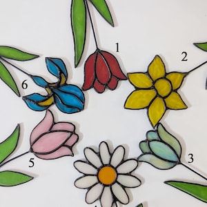 Puede incluir: Una colección de flores de vidrieras en varios colores, incluyendo rojo, amarillo, azul, rosa y blanco. Cada flor tiene un tallo negro y hojas verdes. Las flores están dispuestas en un patrón circular.