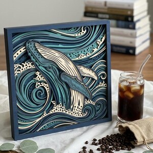 May include: A square, multi-layered wooden wall art piece featuring a humpback whale breaching through stylized ocean waves. The artwork uses a color palette of deep navy, teal, and cream, with intricate cutouts creating a three-dimensional effect.