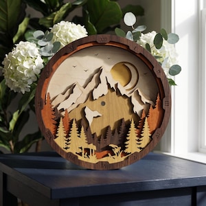 Könnte beinhalten: Eine kreisförmige Wanduhr aus Holz mit einer mehrschichtigen, lasergeschnittenen Berglandschaft, Kiefern, einer Mondsichel und Vögeln. Der dunkle Holzrahmen zeigt römische Ziffern von I bis XII, die um den Rand des Zifferblatts eingraviert sind.
