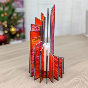 Könnte beinhalten: Ein dekorativer Kerzenhalter mit einzigartigem Design. Er verfügt über eine Reihe rechteckiger Glasmalereien in Rot-, Orange- und Lilatönen, die in einer gebogenen Form angeordnet sind. In der Mitte befindet sich eine weiße Kerze. Der Halter hat einen silberfarbenen Metallrahmen.