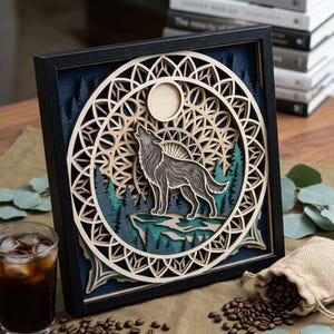 May include: A square black-framed wall art piece featuring a multi-layered laser-cut wood design. The artwork depicts a gray wolf howling at a full moon, set against a backdrop of dark blue pine trees and an intricate geometric circular pattern.