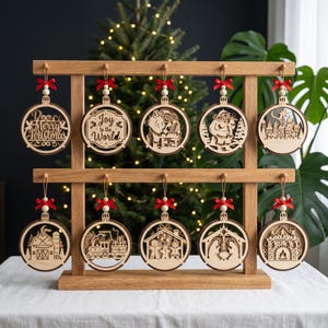 Puede incluir: Un soporte de madera con diez adornos circulares cortados a láser. Cada adorno presenta una escena navideña única, incluyendo textos como Merry Christmas y Joy to the World, decorados con una cuenta de madera y un lazo rojo.