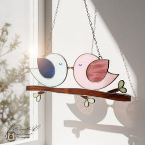 May include: A stained glass artwork depicting two birds kissing on a brown branch. One bird is white with blue detailing, the other is pink with maroon. The piece is suspended by a chain, with green leaves and a logo in the bottom left corner.