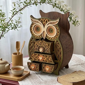 Può includere: Un organizer in legno a forma di gufo composto da più strati, con retro marrone scuro, accenti verde oliva e dettagli color sabbia. Il gufo presenta tre cassetti sovrapposti, ciascuno decorato con intagli a mandala e un piccolo pomello a forma di cuore.