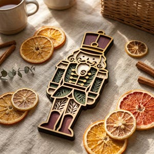 May include: A layered wooden nutcracker ornament featuring a purple hat, a cream-coloured face with a moustache, and a green uniform with gold accents. The ornament is surrounded by dried orange slices and cinnamon sticks on a linen fabric.