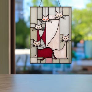 May include: Stained glass panel featuring stylized cats in white, red, and pink. The cats have yellow eyes and black whiskers, set against a clear and light gray background. The panel is framed in black and suspended by a chain.