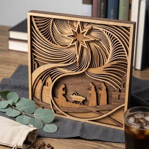 May include: A square, multi-layered wooden wall art piece featuring a laser-cut nativity scene. The design includes silhouettes of figures surrounding a manger, set beneath a large, prominent star with swirling, carved lines in light and dark wood.
