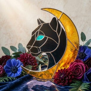 Puede incluir: Obra de arte en vidriera que presenta una cabeza de pantera negra dentro de una luna creciente dorada. La pantera tiene un ojo turquesa. La obra está rodeada de flores rojas oscuras, azules y moradas, y follaje verde, sobre un fondo suave.
