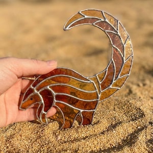 Puede incluir: Una escultura de vidriera de un animal con cola curva, en tonos marrones, naranjas y ámbar. La obra de arte está delineada en plata y descansa sobre una superficie arenosa. La pieza está sostenida por una mano.
