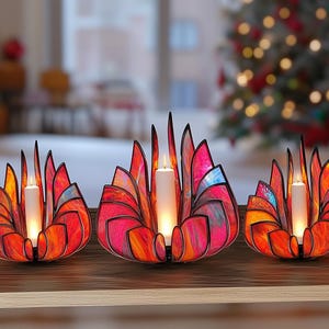 May include: Three decorative candle holders with a stained glass design. The holders are in the shape of a flower, with red, orange, and pink hues. Each holder contains a white candle, and the background is blurred.