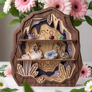 May include: A multi-layered wooden display shelf shaped like a cave, featuring two horizontal shelves holding various raw crystals. The frame is constructed from dark and light wood, with laser-cut crystal motifs and engraved star patterns on the back panel.