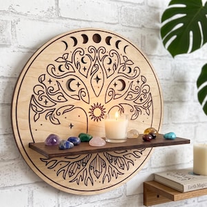 May include: A circular light-colored wooden wall shelf featuring a laser-engraved tree of life design with moon phases, stars, and a sun. A dark brown horizontal shelf is attached to the center, holding a lit white candle and several small crystals.