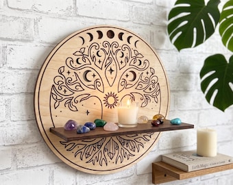 May include: A circular light-colored wooden wall shelf featuring a laser-engraved tree of life design with moon phases, stars, and a sun. A dark brown horizontal shelf is attached to the center, holding a lit white candle and several small crystals.