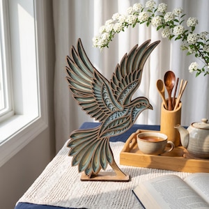 May include: A multi-layered wooden bird sculpture featuring intricate cutouts and a color palette of natural wood tones, soft teal, and cream. The bird is depicted in a dynamic flight pose with wings spread wide, supported by a flat wooden base.