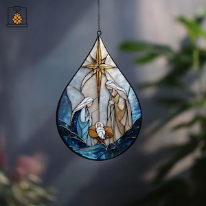 May include: Stained glass teardrop ornament depicting the Nativity scene. The design features Mary, Joseph, and baby Jesus, with a golden star and blue and white accents. The ornament hangs from a chain.