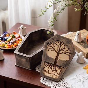 May include: A dark brown wooden coffin-shaped box with a removable lid. The lid features a laser-cut design of a tree with visible roots and a small skeleton figure lying beneath the roots, set against a light-colored wood background.