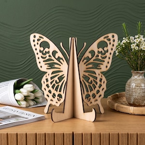 May include: A light-colored wooden display stand featuring a laser-cut butterfly silhouette with intricate wing patterns. The structure consists of two interlocking pieces that form a central support base, designed to hold items upright.