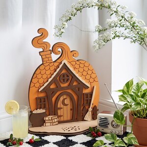 May include: A layered wooden cutout shaped like a pumpkin house with a chimney, a front door featuring a crescent moon cutout, windows, a broom, and a cauldron. The design includes brown and orange tones with detailed roof shingle patterns.