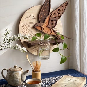 Könnte beinhalten: Ein kreisförmiges Wandkunstobjekt aus Holz mit einem geschichteten Schwalbenmotiv, verziert mit floralen Mustern und Wiener Geflecht. Ein kleines Holzregal trägt ein Glasgefäß mit einer grünen Pflanze vor einem hellen Holzhintergrund.