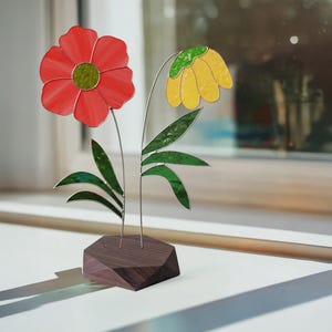 May include: A stained glass floral sculpture. It features a red flower with a green centre and a yellow flower with a green top. The flowers have silver stems and green leaves, set in a dark wooden base. The sculpture is displayed on a windowsill.