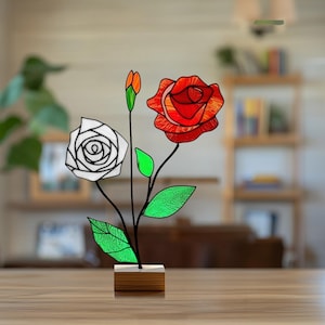 May include: A stained glass rose sculpture featuring a white rose, a red rose, and a bud. The leaves are green, and the stems are black. The sculpture is mounted on a small wooden base. The roses are made of colorful glass.