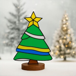 Peut inclure: Un sapin de Noël décoratif avec une étoile jaune au sommet. L'arbre est vert avec des accents blancs, jaunes et bleus. Il repose sur une base ronde en bois marron. L'arrière-plan présente des sapins de Noël flous.