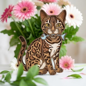 May include: A multi-layered wooden cat figurine featuring a brown and tan leopard-like spotted pattern. The cat is depicted in a sitting position with a light blue fish held in its mouth. The design consists of stacked wood cutouts.
