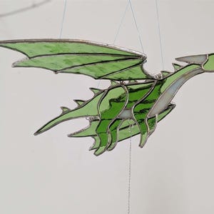 May include: A stained glass dragon sculpture in flight, featuring green wings and body segments. The dragon is suspended by thin wires, showcasing its detailed design and craftsmanship. The wings are a vibrant green, and the body has a segmented design.