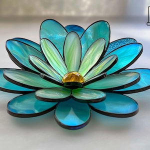 May include: A decorative stained glass flower sculpture. It features turquoise and green petals with a golden centre, all outlined in black. The piece is designed to be a decorative art object.