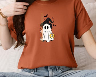 May include: Rust-colored t-shirt featuring a cartoon ghost wearing a witch hat and holding a pool cue. The ghost has a nine-ball and is surrounded by bats. The shirt is a casual, unisex style.