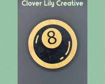 May include: A round wooden coaster featuring a black eight ball design with the number 8 in the center. The coaster has a light brown wooden border and the text "Clover Lily Creative" at the top.