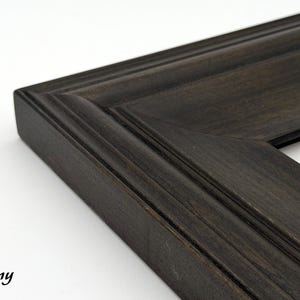 May include: Close-up of a dark ebony-colored picture frame corner. The frame has a beveled edge and a detailed, textured surface. The word "Ebony" is printed in a cursive font in the lower left corner. The frame is likely made of wood.