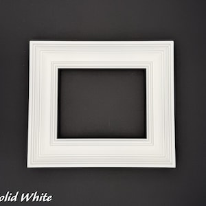 May include: A solid white picture frame with a rectangular opening. The frame has a layered, textured design. The text "Solid White" is at the bottom left corner of the image. The frame is set against a dark background.