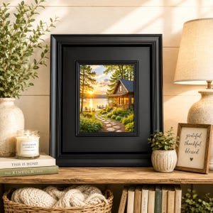 May include: A black picture frame containing a landscape print of a cabin by a lake at sunset, with a stone path leading to the water. The frame is displayed on a wooden shelf alongside a candle, a small potted plant, and a framed sign.