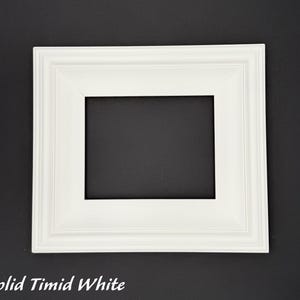 May include: A square, white picture frame with a decorative edge. The frame has a rectangular opening in the center and the text "Solid Timid White" at the bottom. The frame is set against a black background.