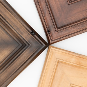 May include: Close-up of four cabinet door samples in different wood finishes: a dark brown, a medium brown, a light brown, and a natural wood finish. The doors are all the same style with raised panels and a simple frame.