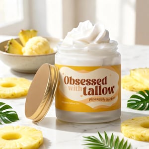Könnte beinhalten: Ein durchsichtiges Glasgefäß gefüllt mit weißem, aufgeschlagenem Talg, verschlossen mit einem goldenen Deckel. Das Etikett zeigt den Text "Obsessed with tallow" und "Pineapple Sorbet" in brauner Schrift auf gelbem Grund.