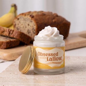 Könnte beinhalten: Ein durchsichtiges Glasgefäß gefüllt mit weißem, aufgeschlagenem Talg, gekrönt von einer Produktspirale. Das Glas hat einen goldenen Deckel und ein gelbes Etikett mit dem braunen Aufdruck "Obsessed with tallow" und "Banana Bread" sowie einer Banane.