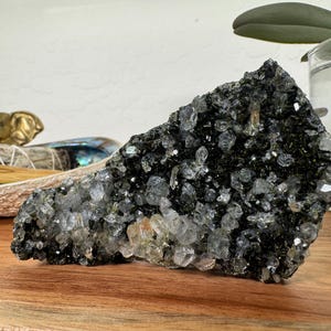 May include: A large, dark gray crystal cluster with clear quartz points. The crystal sits on a wooden surface, with a blurred background featuring a decorative elephant, sage bundles, and a glass container with greenery.