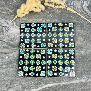 Peut inclure: Ensemble de quatre dessous de verre carrés en céramique noire. Chaque dessous de verre présente un motif floral répété avec des accents blancs, jaunes et turquoise. Les dessous de verre sont disposés sur une surface grise.