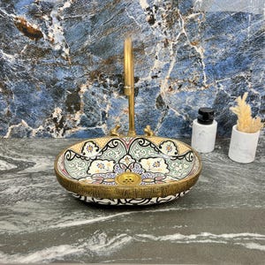 Könnte beinhalten: Ein ovales Keramikwaschbecken mit einem detaillierten Blumenmuster in Grün, Gelb und Rot, eingefasst von einem goldfarbenen Rand. Über dem Waschbecken ist ein goldfarbener Wasserhahn montiert. Zwei kleine weiße Behälter stehen auf der Theke.