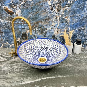 Puede incluir: Un lavabo de cerámica con estampado azul y blanco con un desagüe y un grifo dorados. El lavabo tiene un borde azul y un diseño geométrico detallado. El grifo es curvo y tiene un mango en forma de cruz. El lavabo está sobre una encimera gris.