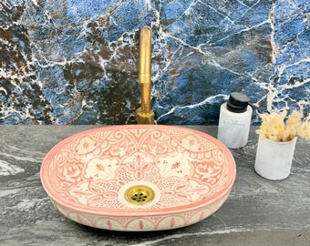 Può includere: Lavandino ovale in ceramica con motivo floreale rosa e bianco, con scarico e rubinetto dorati. Il lavandino è accompagnato da un dosatore di sapone e un piccolo vaso.