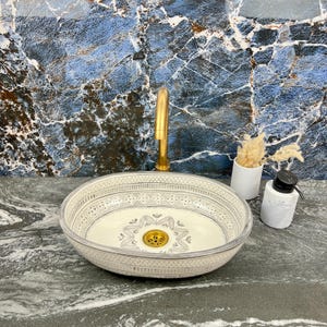 Puede incluir: Un lavabo ovalado de cerámica con un patrón decorativo gris y blanco. Un grifo dorado está montado encima del lavabo. Un jarrón blanco con flores secas y un dispensador de jabón de mármol están en la encimera.