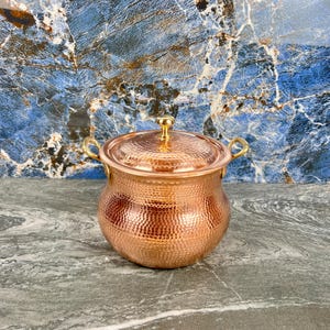 Peut inclure: Une marmite de couleur cuivre avec une texture martelée, un couvercle et deux petites poignées. La marmite a une forme ronde et un bouton doré sur le couvercle. Le fond est un motif de marbre bleu et blanc.