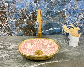 May include: An oval ceramic sink with a pink and white floral design, set within a gold-coloured rim. A gold tap is mounted above the sink. The sink rests on a grey countertop, with a blue and white patterned wall behind.