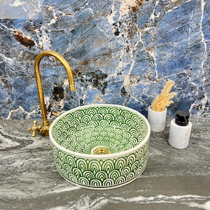 Puede incluir: Un lavabo redondo de cerámica con un diseño verde y blanco, con un grifo de latón. El lavabo está colocado sobre un fondo de mármol azul y gris. También hay un pequeño jarrón blanco con flores secas y un dispensador de jabón de mármol.
