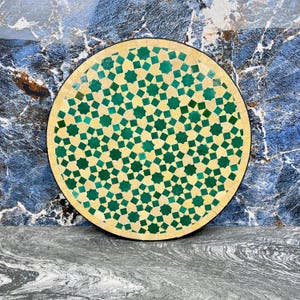 Puede incluir: Mesa redonda de mosaico con fondo crema y un patrón geométrico de estrellas verdes. La mesa tiene un borde negro y está sobre una superficie de mármol azul y gris. El diámetro de la mesa es de aproximadamente 61 cm.