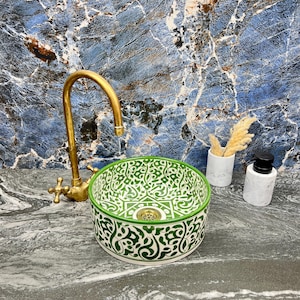 Könnte beinhalten: Ein dekoratives Keramikwaschbecken mit einem grün-weißen Zierornament. Das Waschbecken ist rund mit einem grünen Rand und einem goldfarbenen Abfluss. Ein goldener Wasserhahn befindet sich hinter dem Waschbecken. Ein Marmor-Seifenspender und eine Vase mit getrockneten Pflanzen stehen daneben.