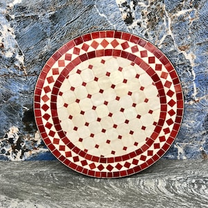 May include: Circular mosaic table with a red and white geometric design. The tabletop has a central pattern of white octagons and red squares, encircled by rings of red diamonds and squares. The table has a black border.